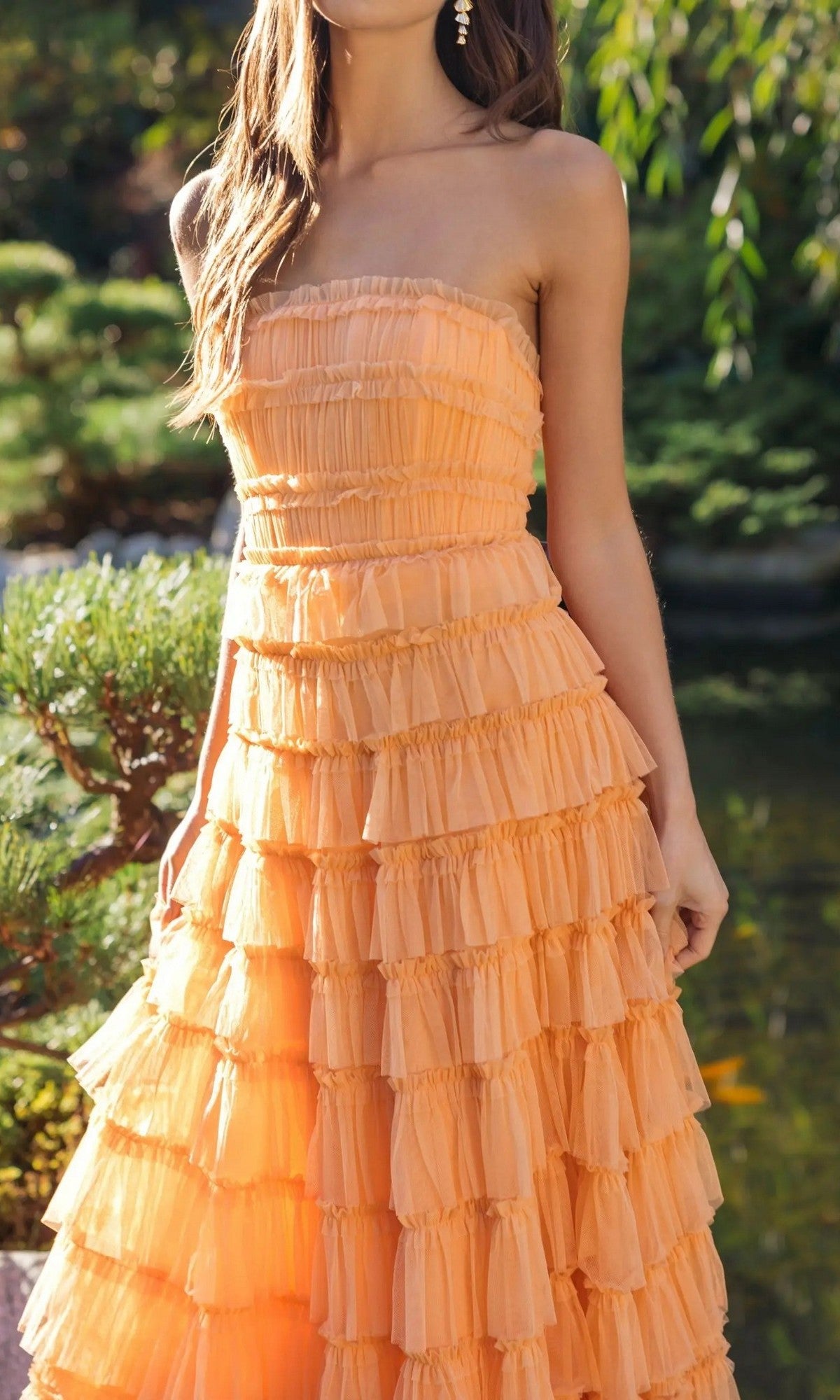 A woman in the Mac Duggal 8286 Midi Semi-Formal Dress, featuring a strapless design and micro-ruffle details, stands outdoors near greenery and water.